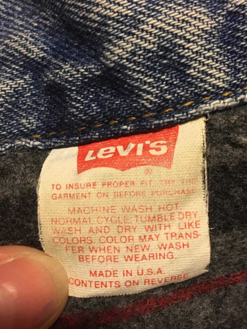 Pode incluir: Um r&oacute;tulo de cuidados Levi's branco com texto vermelho. O r&oacute;tulo tem instru&ccedil;&otilde;es para lavar e secar a roupa. O texto diz: "Para garantir um ajuste adequado, experimente esta roupa antes de comprar. Lavar na m&aacute;quina com &aacute;gua quente. Ciclo normal. Secar na m&aacute;quina a baixa temperatura. Lavar e secar com cores semelhantes. A cor pode transferir ao lavar pela primeira vez. Lavar antes de usar. Feito nos EUA. Conte&uacute;do no verso."