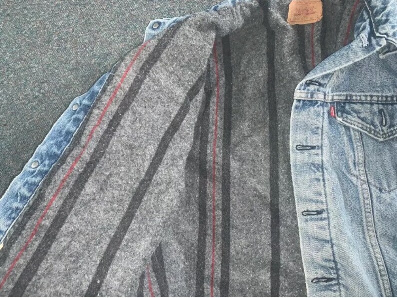 Pode incluir: Uma jaqueta de ganga azul com forro listrado cinza e vermelho. A jaqueta tem fecho de bot&otilde;es e v&aacute;rios bolsos.