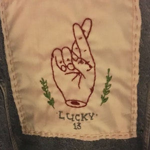 Pode incluir: Um remendo de tecido branco com bordado vermelho de uma m&atilde;o com os dedos cruzados. O remendo &eacute; costurado em uma jaqueta jeans azul. O texto "LUCKY 13" &eacute; bordado abaixo da m&atilde;o.