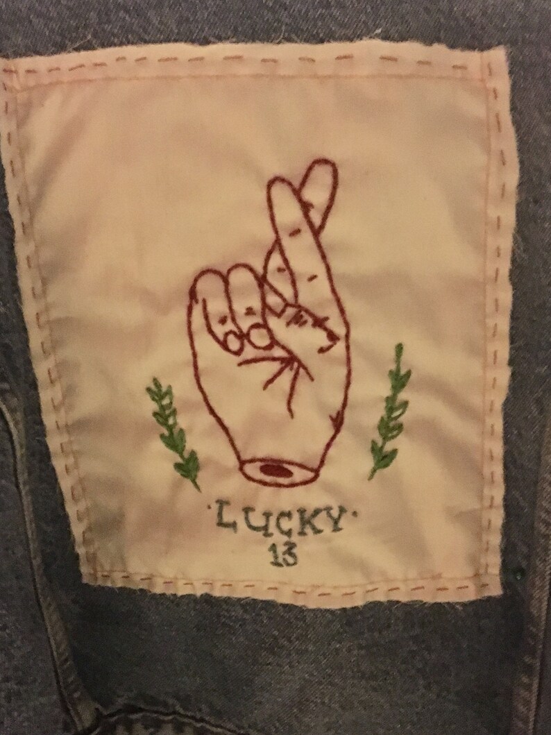 Pode incluir: Um remendo de tecido branco com bordado vermelho de uma m&atilde;o com os dedos cruzados. O remendo &eacute; costurado em uma jaqueta jeans azul. O texto "LUCKY 13" &eacute; bordado abaixo da m&atilde;o.