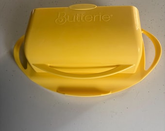 May include: A bright yellow butter dish with the word "Butterie" printed on the lid. The dish has a rectangular lid with a small handle and a rounded base with handles on either side.