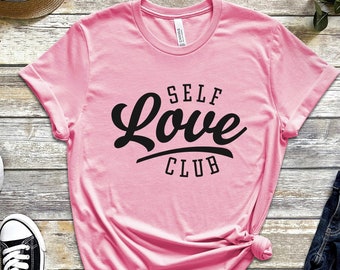 May include: Pink t-shirt with black text that reads "SELF Love CLUB". The shirt is laid flat on a wooden surface with a pair of black sneakers, a straw hat, a watch, and a pair of jeans.