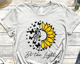 May include: Gray t-shirt with a black and yellow sunflower and butterflies graphic. The text "Be the Light" is printed below the graphic.