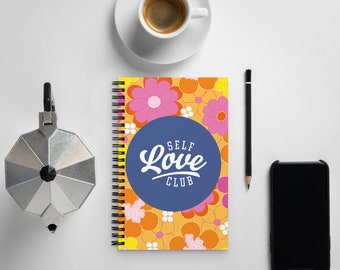 May include: A spiral bound notebook with a yellow background and a colorful floral pattern. The notebook has a blue circle with the text "Self Love Club" in white.