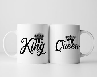 May include: Two white ceramic mugs with black text and crown designs. The mug on the left says "The King" and the mug on the right says "The Queen".