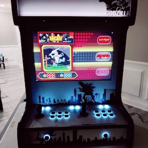 May include: Black arcade machine with a Godzilla theme. The screen displays arcade game options, including "Arcade" and "Classics". The machine has joysticks, buttons, and a cityscape silhouette design. The word "Godzilla" is visible on the top.
