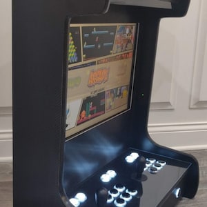 May include: A black arcade cabinet with a screen displaying a variety of classic video games. The cabinet has a carbon fiber finish and white LED lights. The cabinet has a joystick and buttons for playing games.