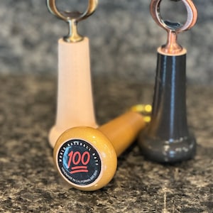 May include: Three wooden bottle openers shaped like baseball bats. One is natural wood, one is black, and one is gold with a black sticker that says "World Baseball Bats 100 Handmade in California Est. 2014".