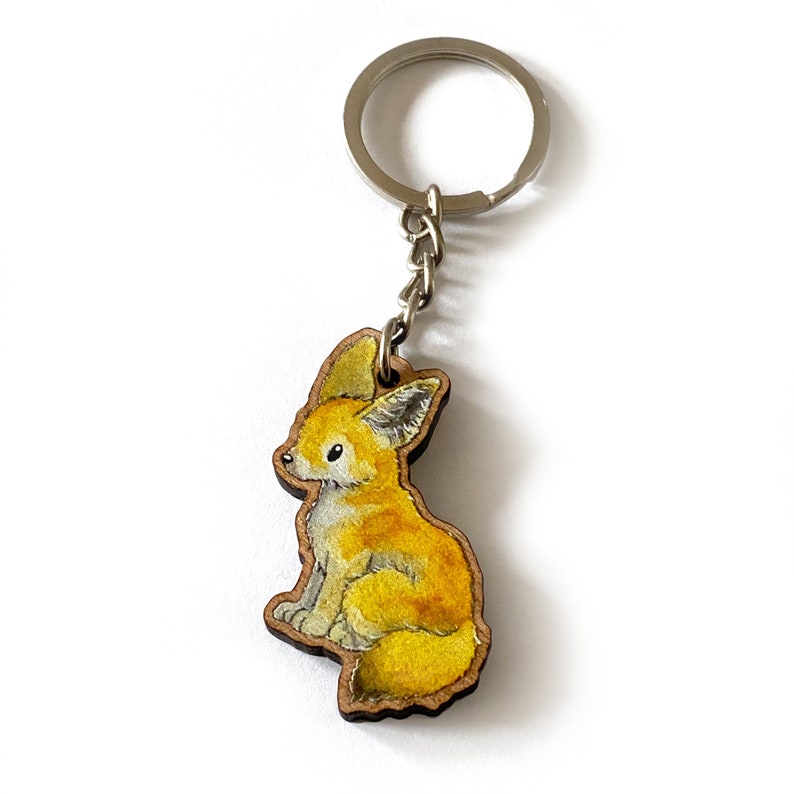 Op de afbeelding: Een houten sleutelhanger met een handgeschilderde illustratie van een fennekvos. De vos zit met zijn oren omhoog en heeft een gele en witte vacht.