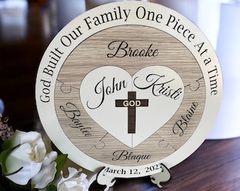 Op de afbeelding: Een houten puzzel in de vorm van een hart met de woorden "God Built Our Family One Piece At A Time" eromheen. Het hart is verdeeld in vijf stukken, elk met een naam: Brooke, John, Kristi, Baylee en Blaine. In het midden van het hart staat een kruis met het woord "God" erop. De datum "March 12, 2023" staat onder het hart.
