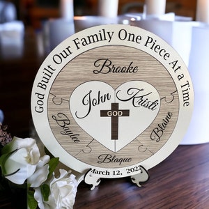 May include: A wooden heart-shaped puzzle with the words "God Built Our Family One Piece At A Time" around the outside. The heart is divided into five pieces, each with a name: Brooke, John, Kristi, Baylee, and Blaine. The center of the heart has a cross with the word "God" on it. The date "March 12, 2023" is written below the heart.