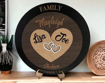 Op de afbeelding: Een ronde houten plaquette met een zwarte rand, met een hartvormige puzzel met de namen "Lisa" en "Joe" erin. De tekst "FAMILY Mayleigh Jackson NOVEMBER 7, 2020" is onder het hart gegraveerd.