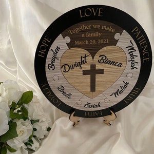 May include: A circular wooden plaque featuring a puzzle piece design with the text "Together we make a family March 20, 2021". The center displays a heart with a cross, names "Dwight" and "Bianca", and surrounding names "Brayden", "Makyah", "Maliah", "Zariah", and "Brielle". The outer black border includes the words "LOVE", "PATIENCE", "TRUST", "FAITH", "LOYALTY", and "HOPE".