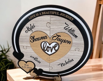 Op de afbeelding: Een houten puzzel in de vorm van een hart met de woorden "Love is a beautiful journey" en de namen "Kylei, Shauna, Jayson, William, Alivia, Nicholas" en de datum "6.27.2020".