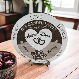 May include: A round wooden family sign with a heart-shaped puzzle design. The sign includes the names "Andrew" and "Dana," the date "Est. July 25, 2020," and the name "Cooper" inside a paw print. The sign is supported by a wooden stand.
