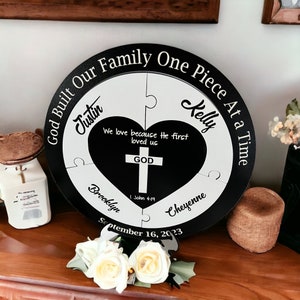 May include: A black and white wooden round sign with a heart in the center. The sign says "God Built Our Family One Piece At a Time". The heart has a cross inside and says "We love because He first loved us. God. 1 John 4:19". The sign also has the names "Justin", "Kelly", "Brooklyn", and "Cheyenne" around the heart. The date "September 16, 2023" is at the bottom of the sign.