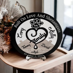 May include: A round wooden family sign with a black border and a white and wood-grain center. The sign features the words "Built On Love And Shenanigans", names, and the date "October 2, 2021". A paw print and heart shape are also included.