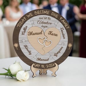 Op de afbeelding: Een houten huwelijkspuzzel met een hartvormig ontwerp, met de namen Manuel en Kara en de datum 31 mei 2026. De puzzel bevat de tekst "Today We Become One Family" en "...and let the Adventure begin..."