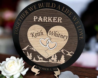 Op de afbeelding: Een ronde houten plaquette met een zwarte rand en een bruine houten kern. De plaquette heeft twee in elkaar grijpende harten met de namen "Keith" en "Whitney" erin. De tekst "Together we build a life we love" is rond de buitenkant van de plaquette aangebracht. De tekst "Parker" bevindt zich boven de harten. De datum "October 5, 2021" bevindt zich onder de harten. De plaquette staat op een houten standaard.