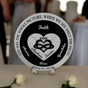 May include: A round, black and white decorative plaque with a heart-shaped puzzle design. The plaque includes the words "Faith", "Love", and names, with the date "June 01, 2024". It is displayed on a white stand.