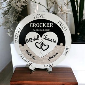 May include: A round wooden plaque with a black and white design. The plaque is divided into two halves, each with a heart-shaped puzzle piece. The top half has the words "LOVE", "CROCKER", "TRUST", and "Est. October 8, 2022". The bottom half has the words "PATIENCE", "LOYALTY", "FAITH", and "HOPE". The heart-shaped puzzle pieces have the names "Mitchell", "Tamara", "Nicholas", and "Amara" written on them.