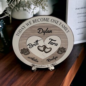 May include: A wooden plaque with a heart-shaped design, featuring the names "Brett" and "Tara" on the left and "Dylan" and "Raelyn" on the right. The plaque also includes the names of two dogs, "Max" and "Laki", and the text "TODAY WE BECOME ONE FAMILY" and "September 3, 2021".