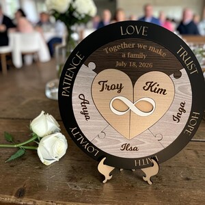 Puede incluir: Una placa de rompecabezas circular de madera con un corazón central y un símbolo de infinito, grabada con los nombres Troy, Kim, Anya, Inga e Ilsa. El texto dice Together we make a family, July 18, 2026, Love, Trust, Hope, Faith, Loyalty y Patience.