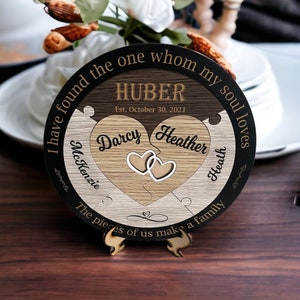 May include: A round wooden sign with a black background and a jigsaw puzzle design. The sign says "I have found the one whom my soul loves" and "HUBER Est. October 30, 2021". The puzzle pieces are shaped like hearts and have the names "Darcy McKenzie" and "Heather Heath" written on them.
