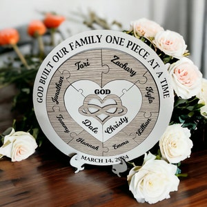 May include: A circular wooden plaque with a heart-shaped cutout in the centre. The plaque is divided into six jigsaw pieces, each with a name inscribed on it. The text "GOD BUILT OUR FAMILY ONE PIECE AT A TIME" is written around the edge of the plaque. The date "MARCH 14, 2024" is written below the plaque.
