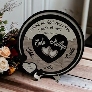May include: A round wooden plaque with a black border and a white background. The plaque has a heart-shaped cutout with two smaller hearts inside. The text "I thank my God every time I think of you. Philippians 1:3" is written in black on the white background. The names "Josh" and "Kaitlyn" are written in black on the heart-shaped cutout. The date "Est 07-10-2021" is written in black on a small heart-shaped cutout at the bottom of the plaque. The word "Ace" is written in black on the wooden background.