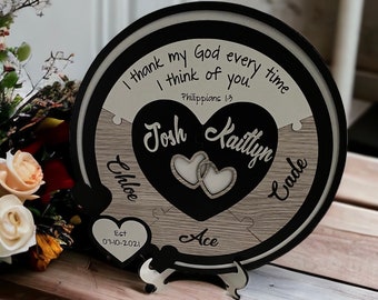 Op de afbeelding: Een ronde houten plaquette met een zwarte rand en een witte achtergrond. De plaquette heeft een hartvormige uitsparing met twee kleinere hartjes erin. De tekst "I thank my God every time I think of you. Philippians 1:3" is in zwart op de witte achtergrond geschreven. De namen "Josh" en "Kaitlyn" zijn in zwart op de hartvormige uitsparing geschreven. De datum "Est 07-10-2021" is in zwart op een klein hartvormig uitsparing aan de onderkant van de plaquette geschreven. Het woord "Ace" is in zwart op de houten achtergrond geschreven.