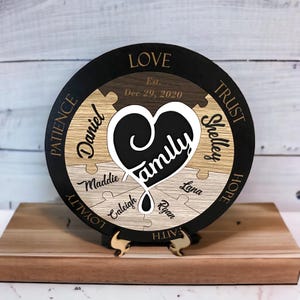 May include: A round wooden plaque with a black background and a heart-shaped puzzle piece in the center. The heart is surrounded by puzzle pieces with the words "Love", "Trust", "Patience", "Hope", "Loyalty", "Faith" and the names "Daniel", "Shelley", "Maddie", "Caleigh", "Lana", "Ryan". The plaque also includes the text "Est. Dec 29, 2020".