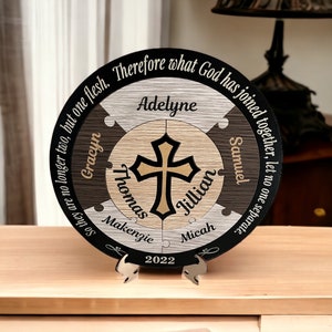 May include: A wooden plaque with a cross in the center and the names Adelyne, Gracyn, Thomas, Jillian, Samuel, Makenzie, and Micah around it. The plaque also includes the quote "So they are no longer two, but one flesh. Therefore what God has joined together, let no one separate." and the year 2022.