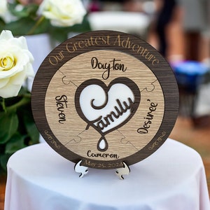 May include: A wooden round puzzle piece with a heart in the center that says "Family". The puzzle pieces are labeled with the names "Steven", "Dayton", "Desinee", and "Cameron". The date "May 25, 2024" is written below the name "Cameron". The puzzle piece is on a white surface and says "Our Greatest Adventure" around the outside.
