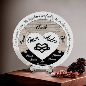 May include: A circular wooden plaque in white, black, and wood tones, featuring a heart-shaped puzzle piece at its centre. The heart is divided into two halves, each inscribed with a name, "Jason" and "Audra." The plaque is encircled by the text "Each special piece fits together perfectly to make our family complete!" The names "Jacob", "Emma", and "Jane" are displayed on the puzzle pieces surrounding the heart. The date "November 12, 2022" is located at the bottom of the plaque. The plaque rests on a wooden surface.
