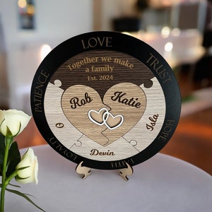 May include: A wooden heart-shaped puzzle with the words "Together we make a family" and "Est. 2024" engraved on it. The puzzle is divided into two halves, each with a heart shape cut out. The names "Rob", "Katie", and "Devin" are engraved on the hearts. The puzzle is surrounded by a black circle with the words "Love", "Trust", "Patience", "Loyalty", "Faith", "Hope", and "Isla" engraved on it.