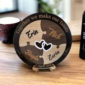 May include: A round wooden plaque with a black border and four puzzle pieces. The puzzle pieces are made of different types of wood and have the names "Erin", "Nick", "Baylin", and "Zaria" written on them. The plaque also has the text "Together we make our family" and "2 April 2022" written on it.