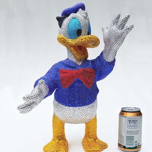 May include: A bedazzled Donald Duck figurine, featuring a blue jacket, red bow tie, and silver gloves. The figure's head and feet are yellow, with a blue hat. A can of soda is next to the figurine.