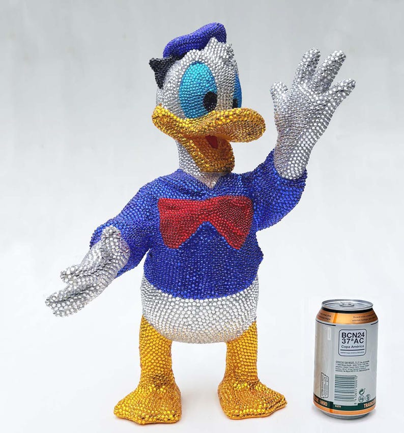 May include: A bedazzled Donald Duck figurine, featuring a blue jacket, red bow tie, and silver gloves. The figure's head and feet are yellow, with a blue hat. A can of soda is next to the figurine.