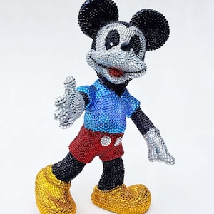 May include: A bedazzled Mickey Mouse figurine. The figure has black ears, arms, and legs, a silver face and hands, a blue shirt, red shorts, and yellow shoes. The figurine is posed with one arm extended.