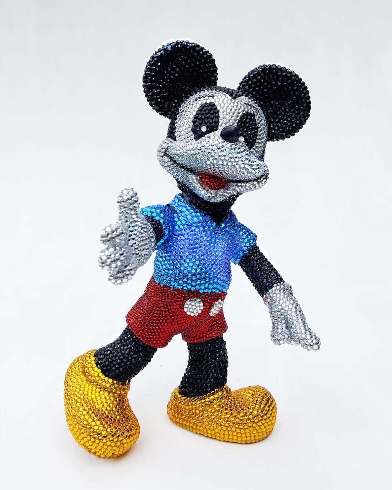 May include: A bedazzled Mickey Mouse figurine. The figure has black ears, arms, and legs, a silver face and hands, a blue shirt, red shorts, and yellow shoes. The figurine is posed with one arm extended.