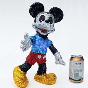 May include: A bedazzled Mickey Mouse figurine in a welcoming pose. The figure is covered in sparkling crystals, with a blue shirt, red shorts, and yellow shoes. A can of soda is next to the figurine.