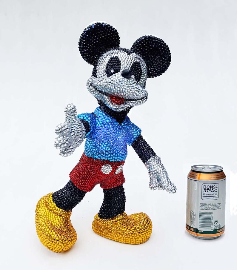 May include: A bedazzled Mickey Mouse figurine in a welcoming pose. The figure is covered in sparkling crystals, with a blue shirt, red shorts, and yellow shoes. A can of soda is next to the figurine.
