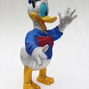 May include: A bedazzled Donald Duck figurine. The figure is wearing a blue sailor shirt, a red bow tie, and a blue hat. The beak and feet are gold, and the eyes are blue. The figure is covered in silver and gold rhinestones.