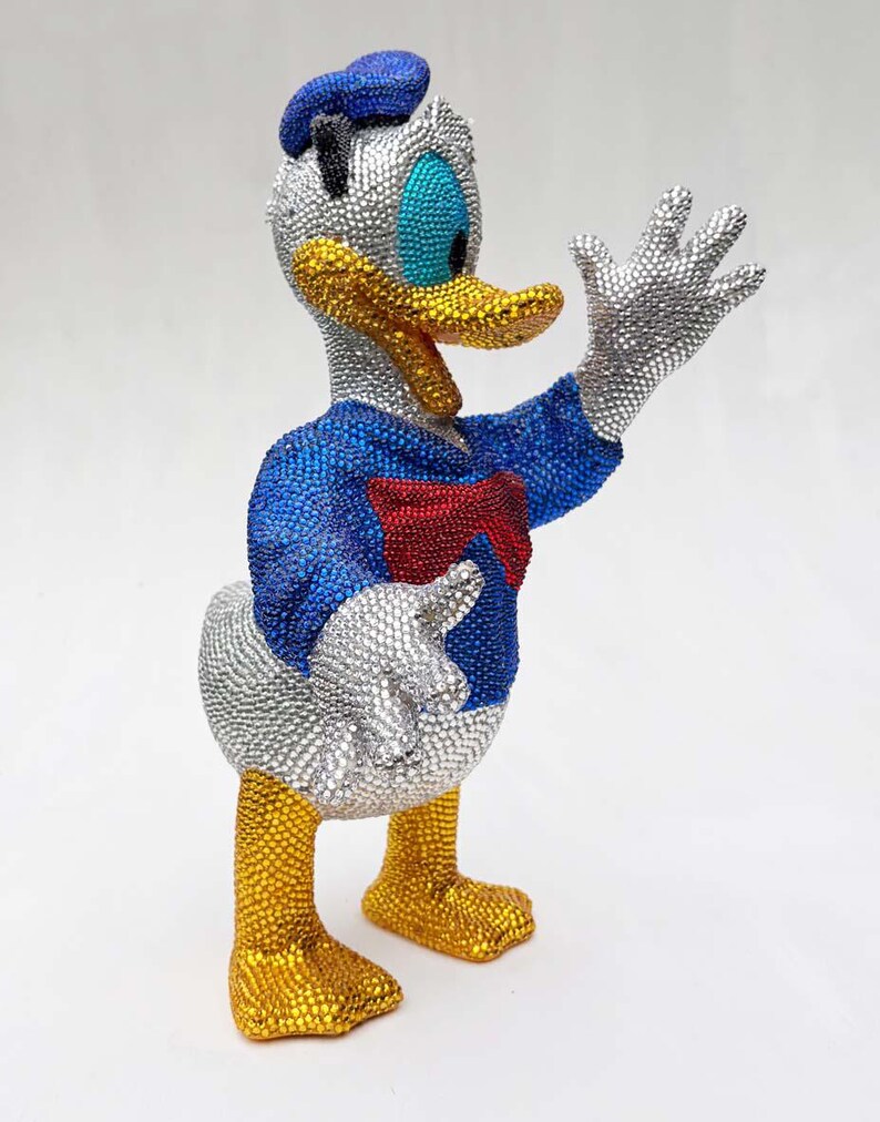 May include: A bedazzled Donald Duck figurine. The figure is wearing a blue sailor shirt, a red bow tie, and a blue hat. The beak and feet are gold, and the eyes are blue. The figure is covered in silver and gold rhinestones.