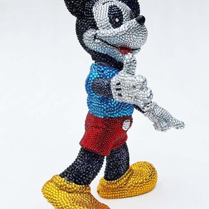 May include: A bedazzled Mickey Mouse figurine. The figure is covered in sparkling rhinestones, with a black head and body, silver face and hands, red shorts, blue shirt, and yellow shoes. The figure is in a walking pose.
