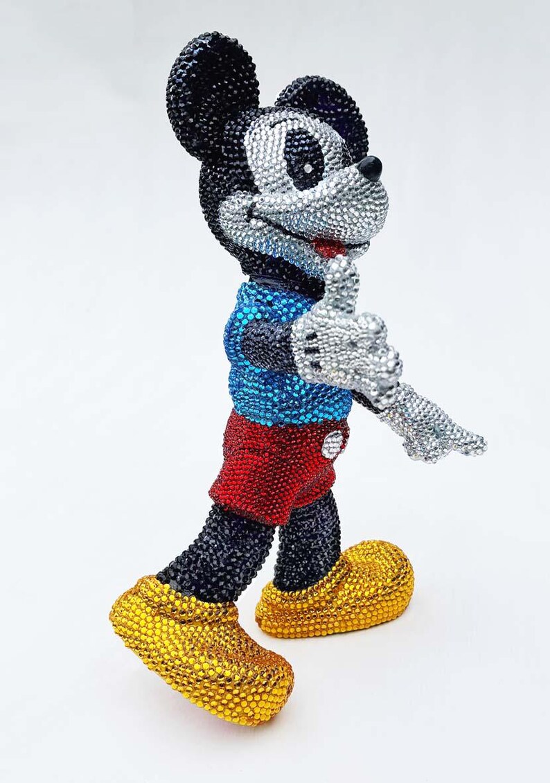 May include: A bedazzled Mickey Mouse figurine. The figure is covered in sparkling rhinestones, with a black head and body, silver face and hands, red shorts, blue shirt, and yellow shoes. The figure is in a walking pose.