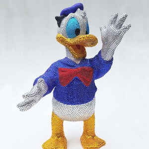 May include: A bedazzled Donald Duck figurine. The figure is covered in sparkling rhinestones, with a blue jacket, red bow tie, and yellow feet. The hat and gloves are silver, and the beak is yellow. The figurine is posed with one hand raised.