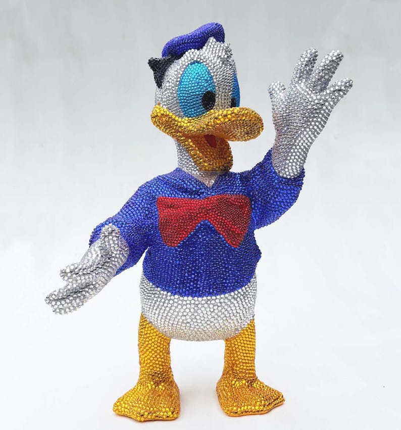 May include: A bedazzled Donald Duck figurine. The figure is covered in sparkling rhinestones, with a blue jacket, red bow tie, and yellow feet. The hat and gloves are silver, and the beak is yellow. The figurine is posed with one hand raised.