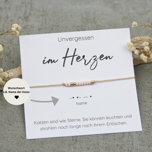 Könnte beinhalten: Eine weiße Karte mit dem Text "Unvergessen im Herzen" und einem kleinen Armband aus Schnur mit rosa und goldenen Perlen. Die Karte enthält auch den Text "Wunschwort z.B. Name der Katze" und ein kleines Herz.